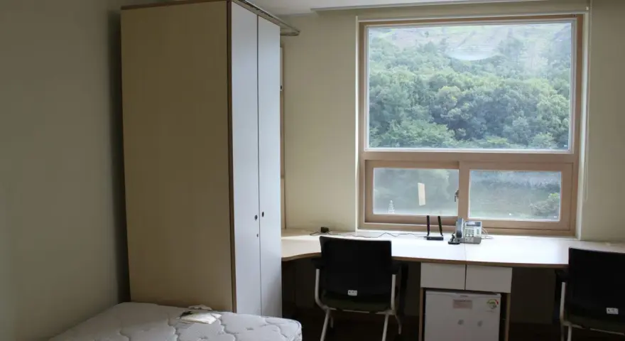 Chambre simple pour un logement étudiant à Séoul, avec un lit, une armoire, un bureau avec mini-frigo et une vue sur la verdure.