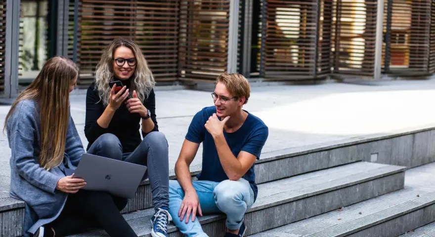 Trois jeunes discutent sur des marches, l'un regarde son téléphone, l'autre un ordinateur portable. Ils semblent parler du cout de la vie aux Pays-Bas.