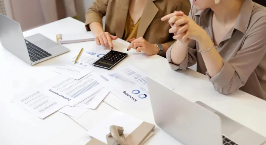 Deux femmes analysent des documents financiers et utilisent une calculatrice. Leurs mains pointent vers des graphiques, suggérant une discussion sur le pilotage de la performance, peut-être dans le cadre d'un msc audit à KEDGE.
