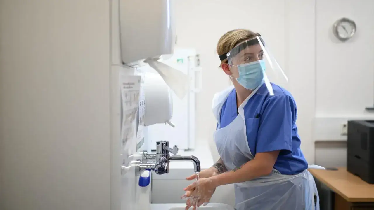 Une infirmière en Espagne, portant masque et visière, se lave les mains sous un robinet.