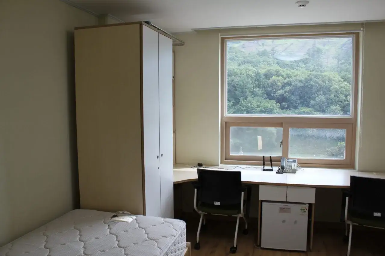 Chambre simple pour un logement étudiant à Séoul, avec un lit, une armoire, un bureau avec mini-frigo et une vue sur la verdure.