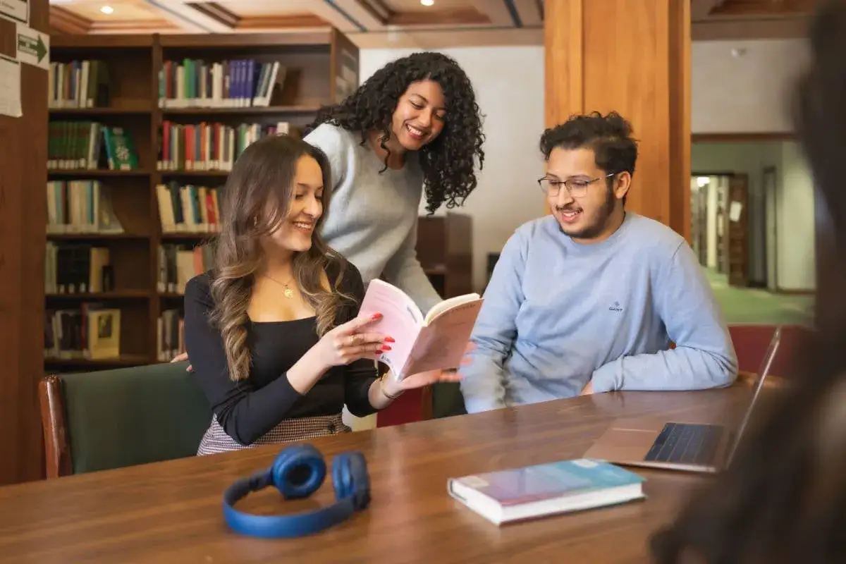 Trois étudiants, dont un master moyen orient, étudient ensemble dans une bibliothèque, partageant un livre et un ordinateur portable.