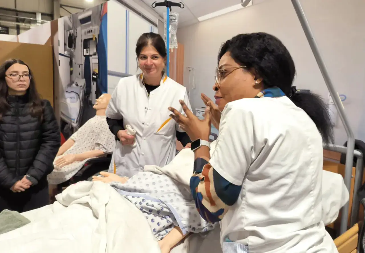 Une aide soignante belge, en formation, explique une procédure à des mannequins dans une salle de simulation médicale.