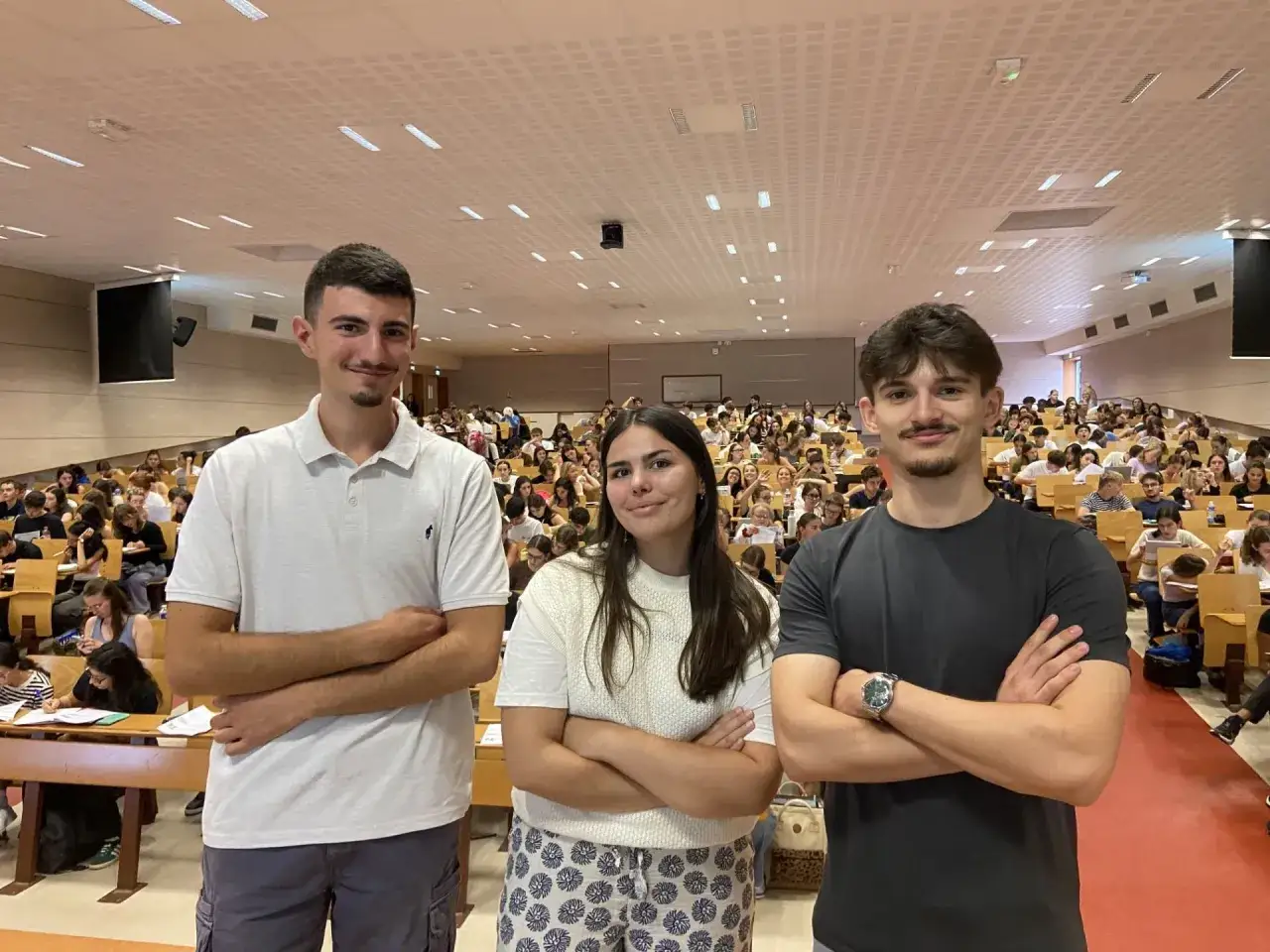 Trois étudiants en médecine posent dans un amphithéâtre rempli d'autres étudiants.