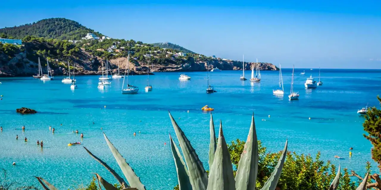Vue idyllique d'une baie aux Baléares, avec des voiliers et des baigneurs. On imagine les étudiants de l'université des îles baléares profiter de ce décor.