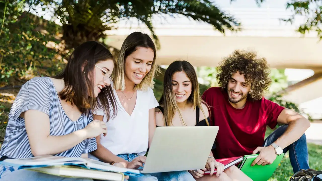 Quatre étudiants souriants travaillent sur un ordinateur portable en plein air, préparant leur master psychologie à l'étranger.