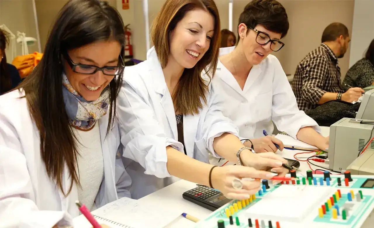 Trois femmes en blouse blanche travaillent sur un circuit électronique, souriantes. Elles suivent une formation accélérée audioprothésiste.
