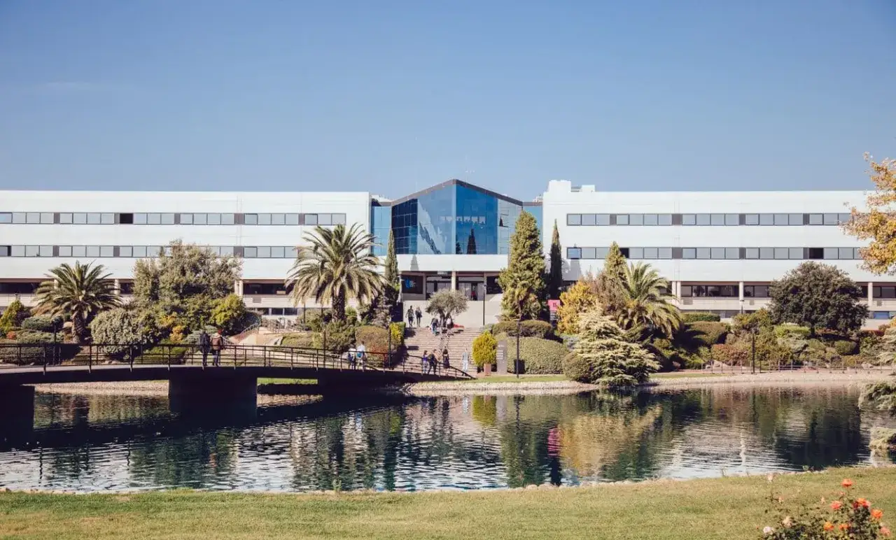 Un campus verdoyant avec un étang paisible et un pont menant à un bâtiment moderne, évoquant une école d'élite à Madrid.