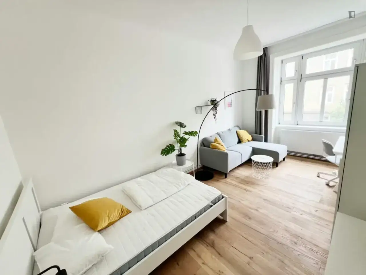Chambre lumineuse pour logement &eacute;tudiant &agrave; Vienne, Autriche. Lit simple, canap&eacute; gris, bureau et grande fen&ecirc;tre.