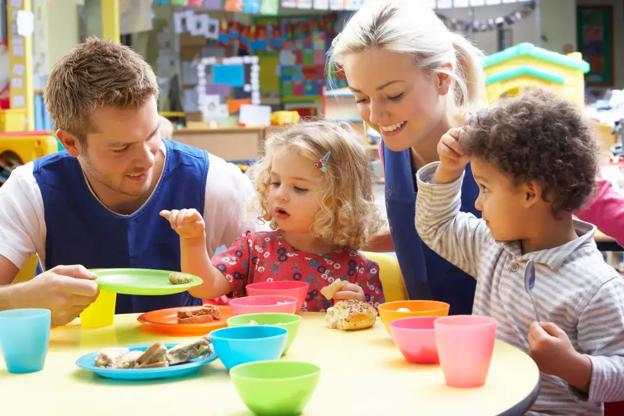 Un homme et une femme souriants, peut-&ecirc;tre en formation de master petite enfance &agrave; distance, partagent un go&ucirc;ter avec deux enfants dans une salle color&eacute;e.