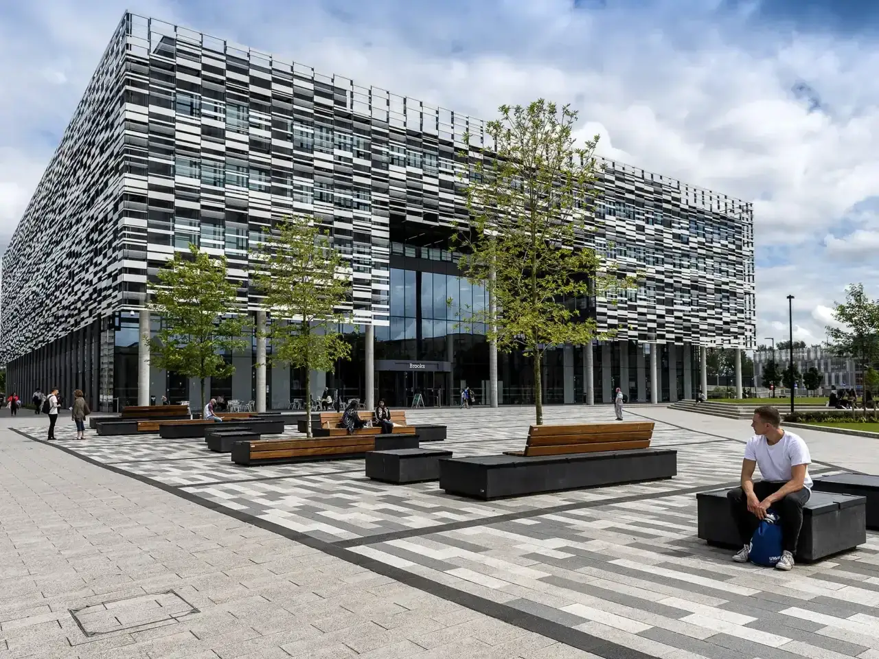 Fa&ccedil;ade moderne de l'universit&eacute; m&eacute;tropolitaine de Manchester. Des &eacute;tudiants profitent du soleil sur des bancs ext&eacute;rieurs.
