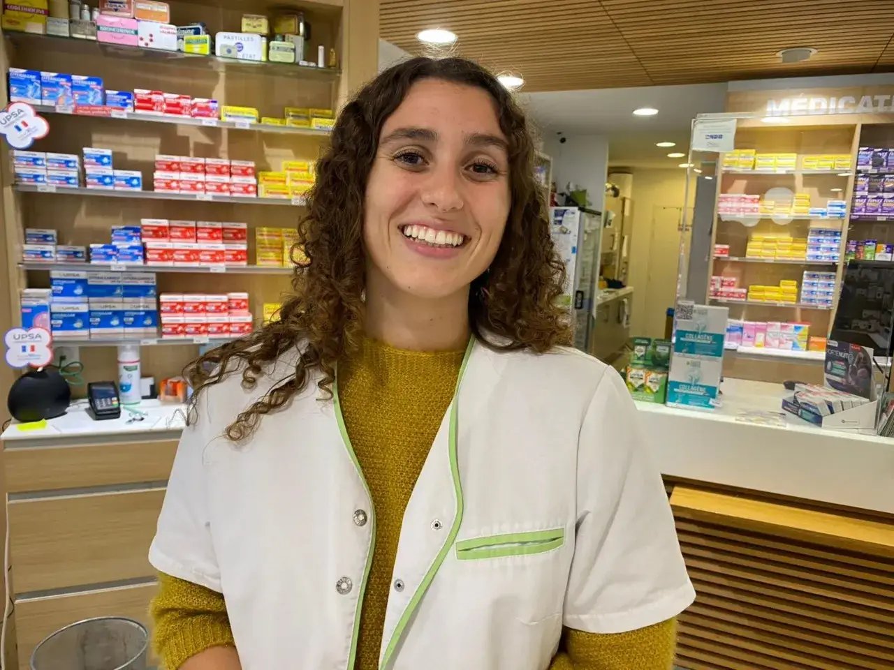 Une souriante pharmacienne travaille dans une pharmacie en Espagne, entourée de boîtes de médicaments colorées.