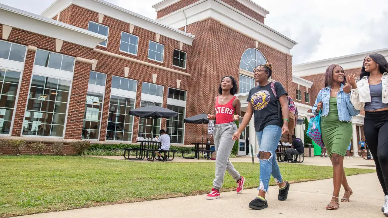 Trois étudiantes marchent sur le campus d'un college aux États-Unis, devant un bâtiment en brique.