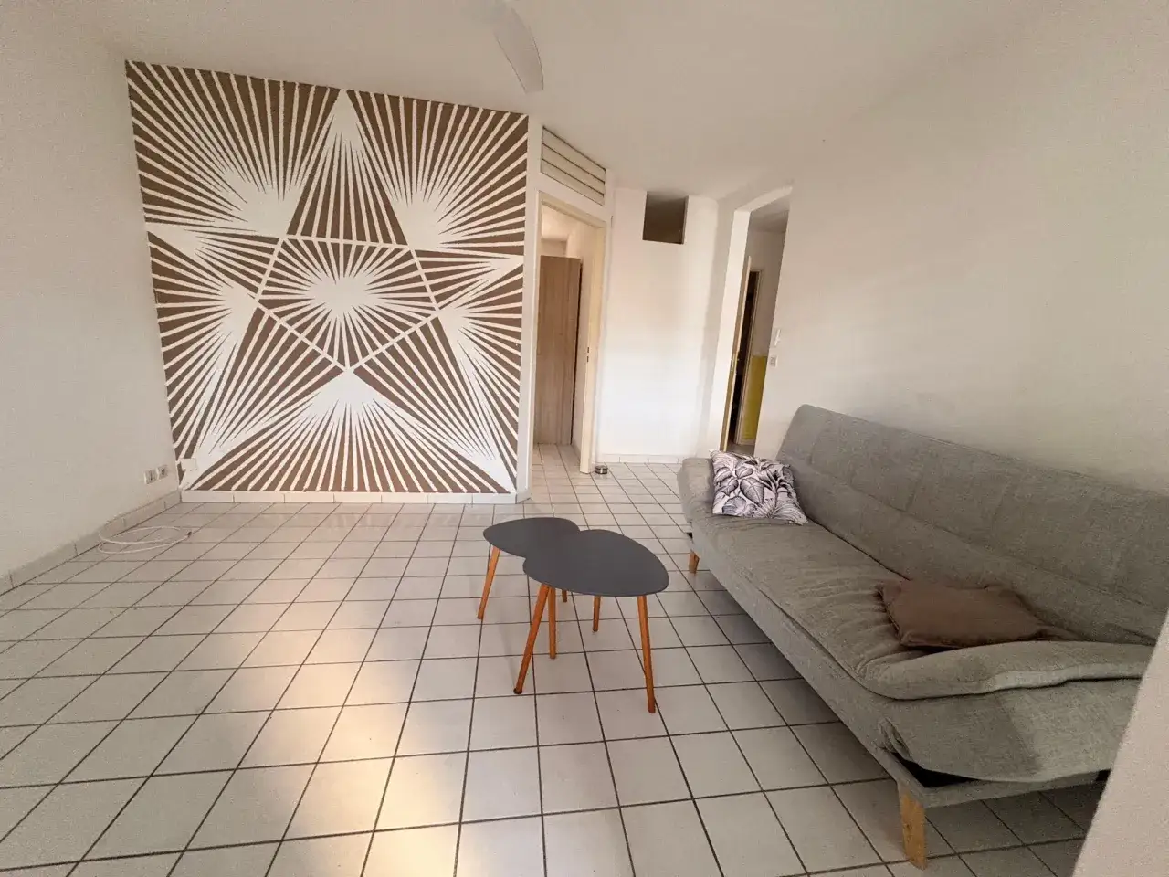 Salon lumineux avec canapé gris, tables basses design et mur décoré d'un motif géométrique. Idéal pour un logement étudiant en Guadeloupe.