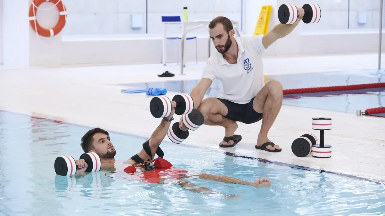 Un étudiant en kinésithérapie à l'école kine Gerone fait des exercices aquatiques avec des haltères, guidé par un professeur.