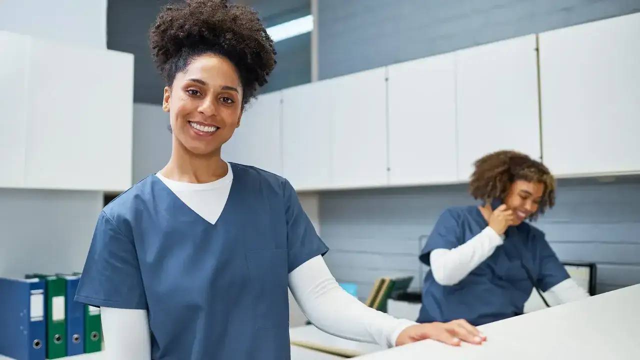 Secrétaire médicale souriante, prête pour sa formation en ligne. Une collègue au téléphone en arrière-plan.