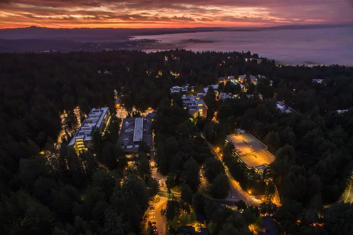 Vue aérienne de nuit sur le campus de UC Santa Cruz, illuminé par des lumières chaudes, avec un ciel orangé et une mer de brouillard au loin.