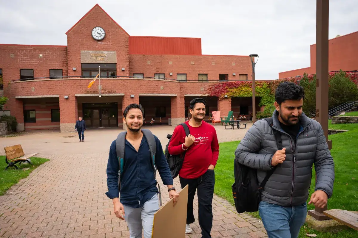 Trois étudiants marchent sur un campus au Canada, portant des sacs à dos. L'un d'eux tient une grande planche en carton.