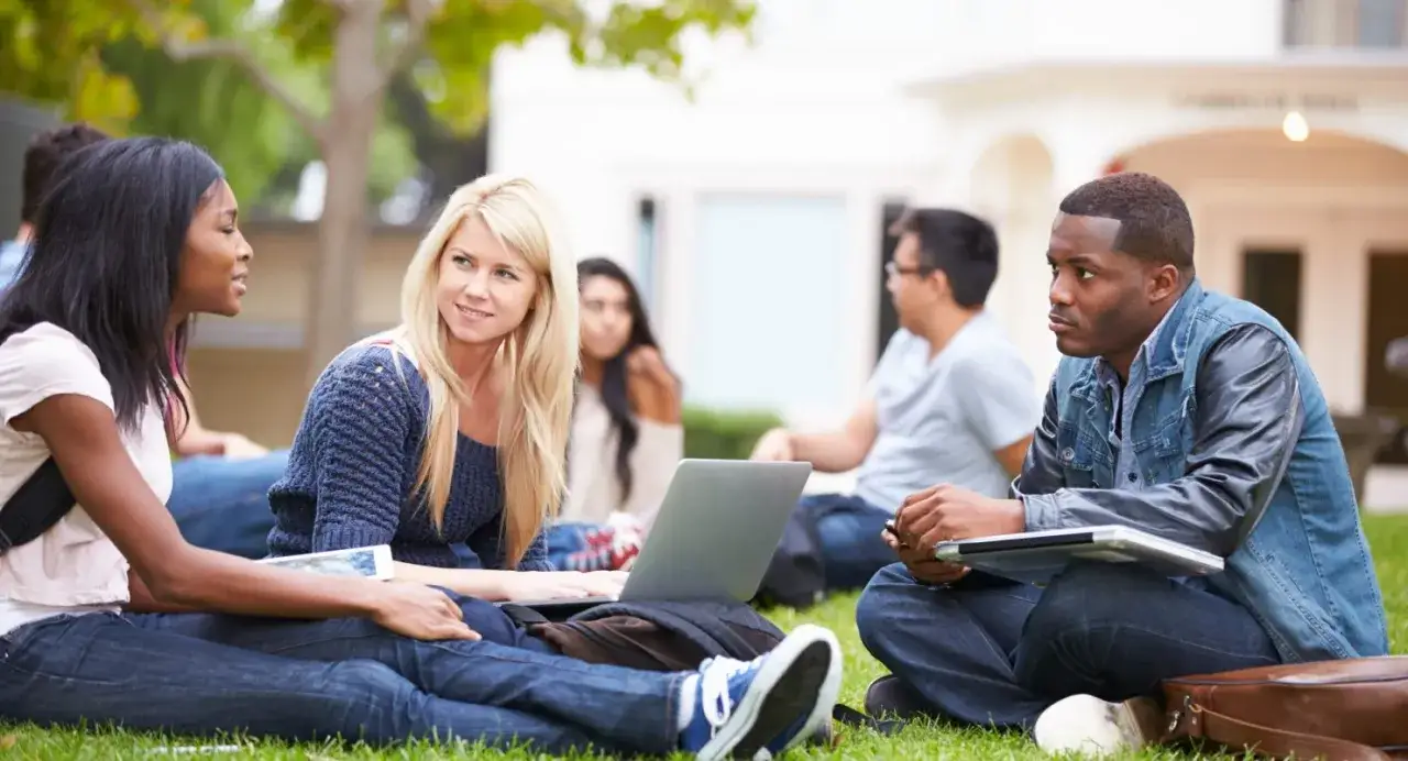 Trois étudiants discutent sur l'herbe, un ordinateur portable ouvert. Ils préparent peut-être leur master en anglais, diplôme en vue.