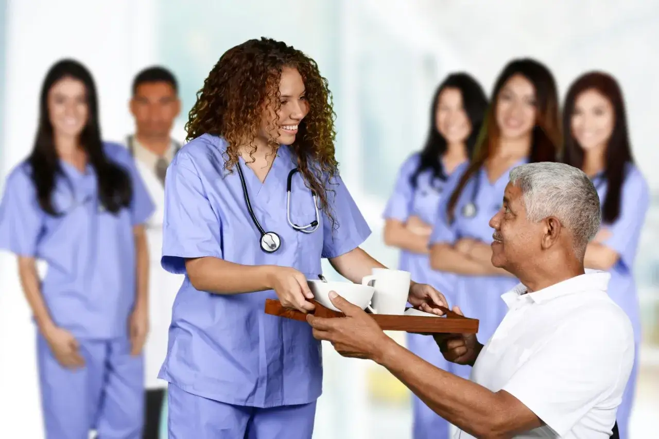 Une jeune femme en formation paramédicale sans bac, souriante, sert un plateau repas à un patient. D'autres soignants sont en arrière-plan.