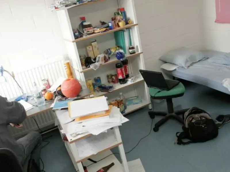 Bureau désordonné, lit simple, étagère remplie d'articles divers. Un ordinateur portable sur une chaise. Une scène typique pour trouver un logement en Belgique.